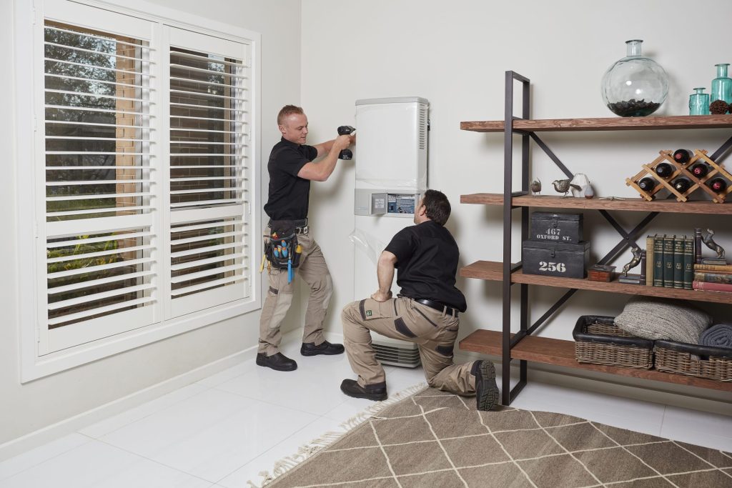 Two tradesmen installing an indoor unit of a heat pump in a modern living room.