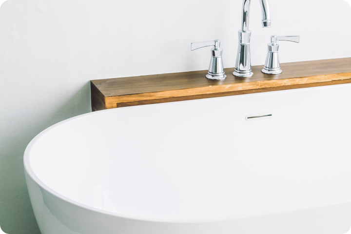 Modern white bathtub with chrome faucets on a wooden shelf against a white wall.