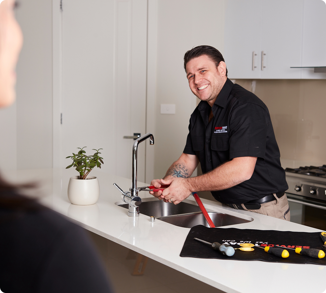 A smiling plumber in a black shirt fixes a kitchen sink faucet with red pliers.