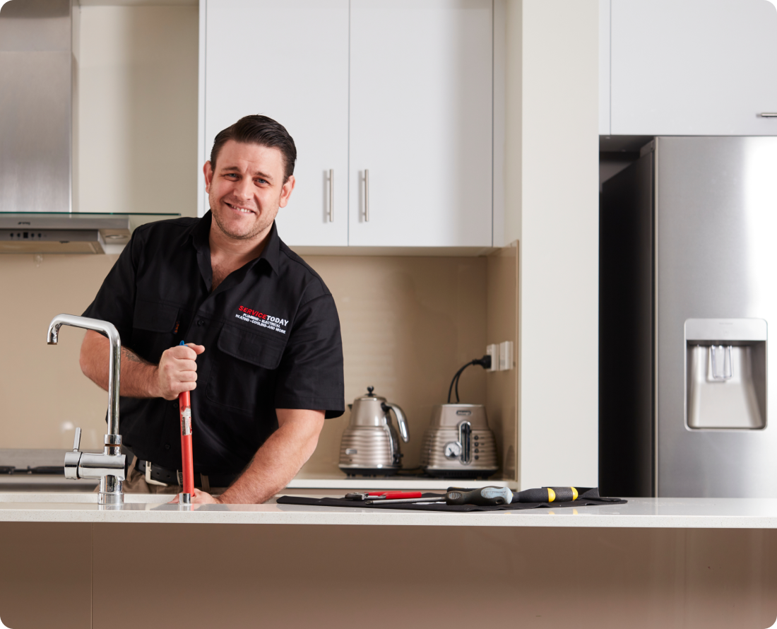 A plumber smiles while using a plunger in a kitchen sink.