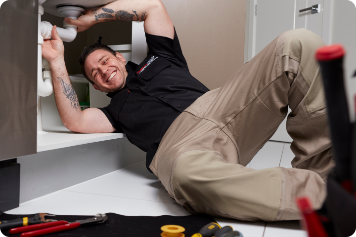 Plumber smiles while working on pipes under a sink with tools nearby.