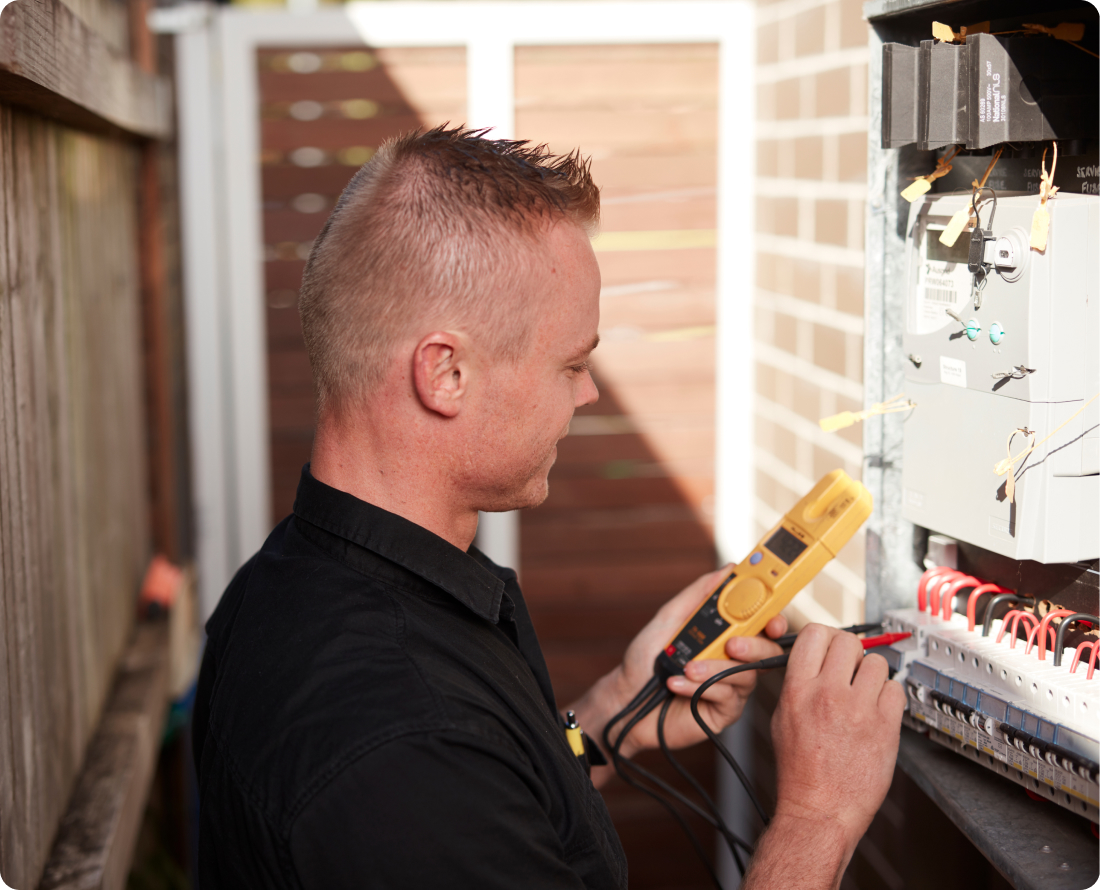 Electrician in black shirt testing electrical panel with yellow multimeter.