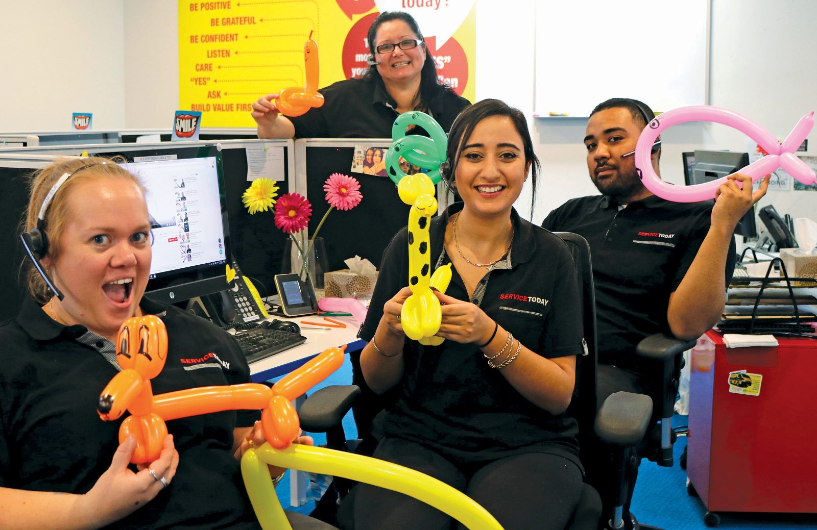 Colleagues at ServiceToday desk playfully holding balloon animals, a giraffe, dog, and fish.