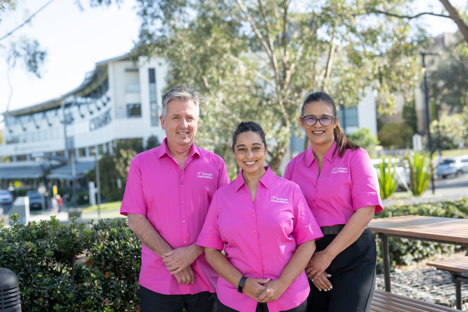 Three people in pink McGrath Foundation shirts smile outdoors in front of a building.